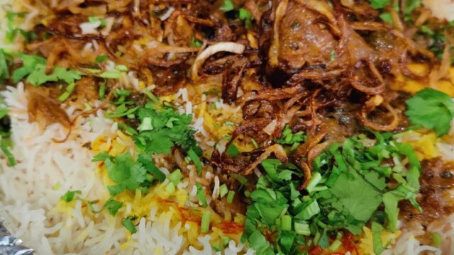 Close-up of a tray of Hyderabadi-style biryani garnished with fried onions and fresh coriander, served in foil container.
