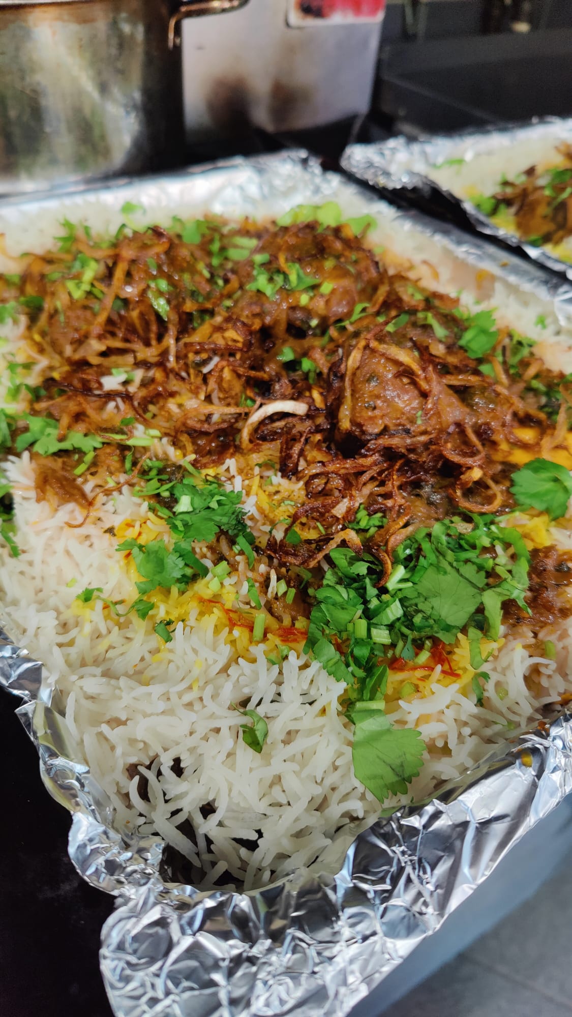 Close-up of a tray of Hyderabadi-style biryani garnished with fried onions and fresh coriander, served in foil container.