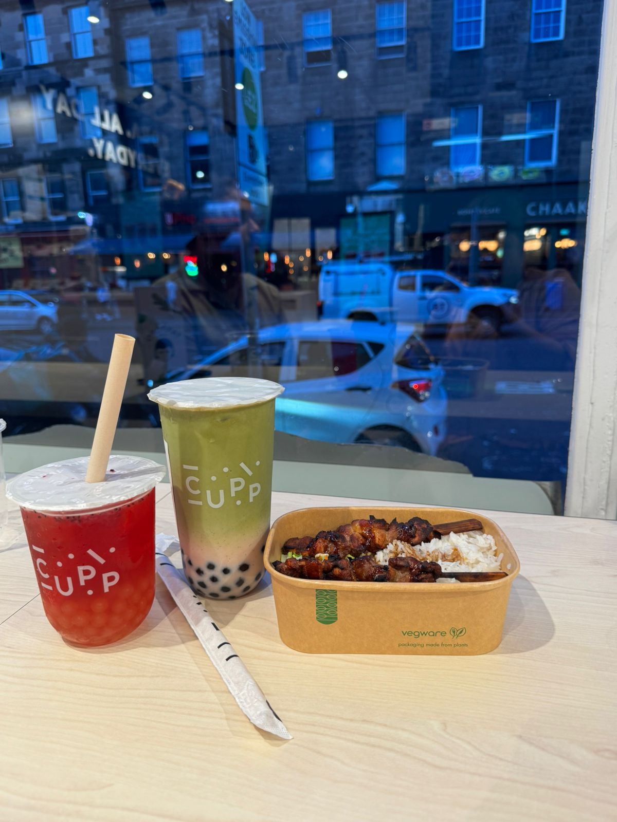 Two bubble teas from CUPP and a rice-and-skewers meal in a compostable Vegware container, placed on a table inside a café with a view of a busy Edinburgh street.