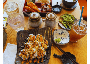 Top-down view of a wooden table at Don@Tokyo, Edinburgh, featuring a red lacquered prawn tempura bento box with rice, gyoza and greens; a black ceramic plate of chicken katsu sushi rolls drizzled with spicy mayo and unagi sauce; a bowl of miso soup with spring onions; a small plate of edamame; two ceramic condiment jars; a glass of iced peach hojicha; and the “Scottish Indian.com” logo banner at the top.