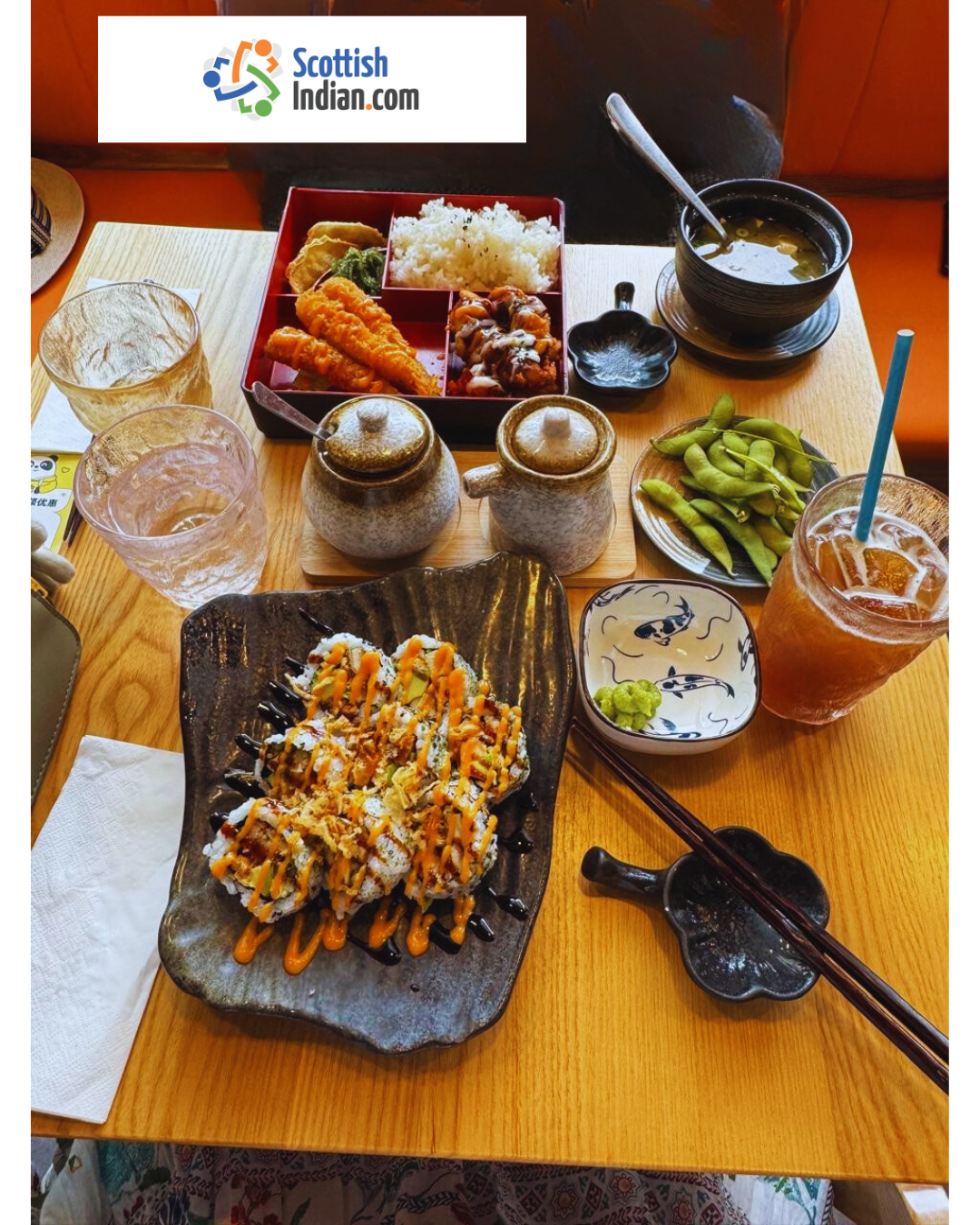 Top-down view of a wooden table at Don@Tokyo, Edinburgh, featuring a red lacquered prawn tempura bento box with rice, gyoza and greens; a black ceramic plate of chicken katsu sushi rolls drizzled with spicy mayo and unagi sauce; a bowl of miso soup with spring onions; a small plate of edamame; two ceramic condiment jars; a glass of iced peach hojicha; and the “Scottish Indian.com” logo banner at the top.