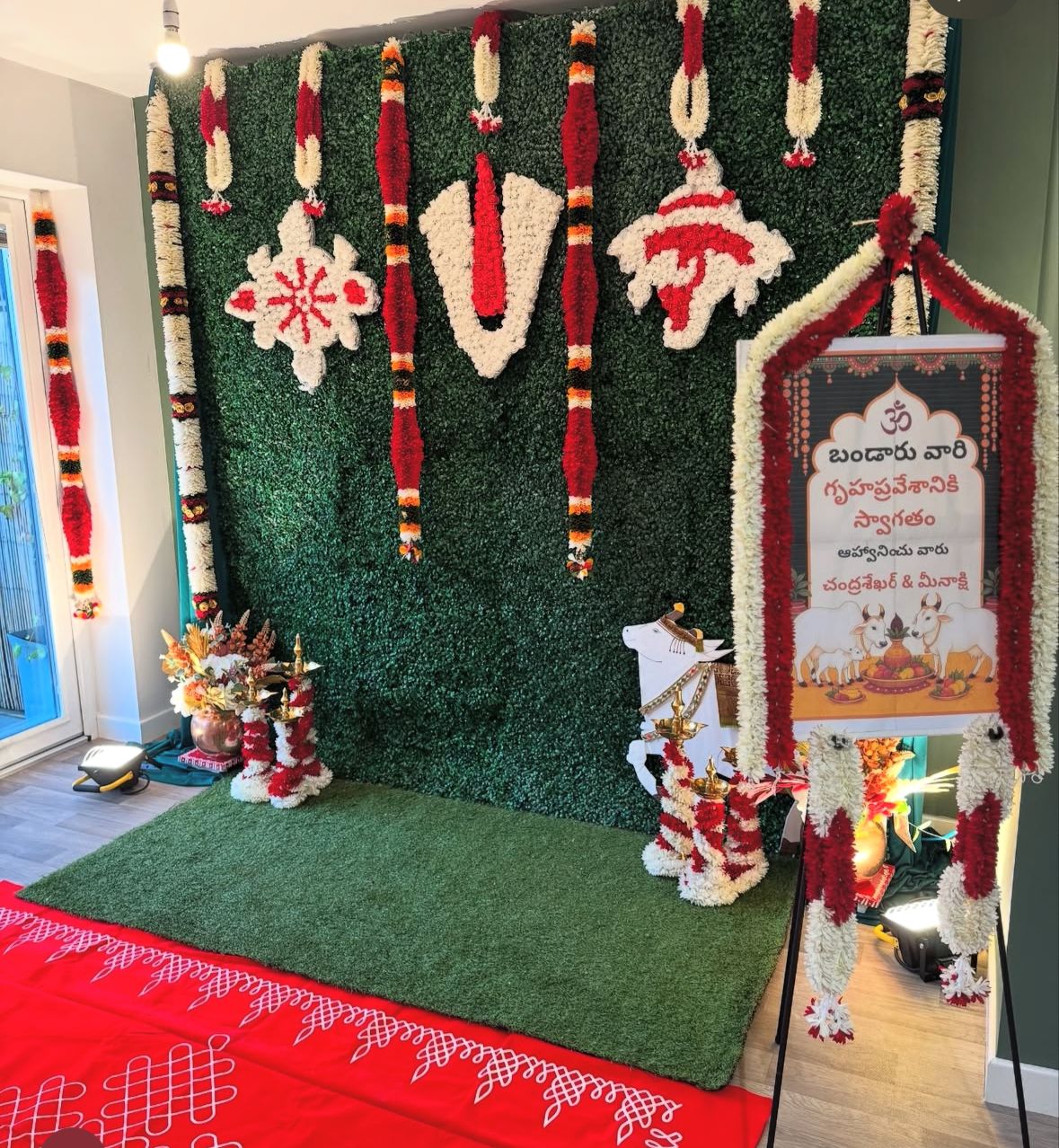 Sacred Vaishnava symbols and red-white flower garlands on a green pooja backdrop with Telugu welcome board